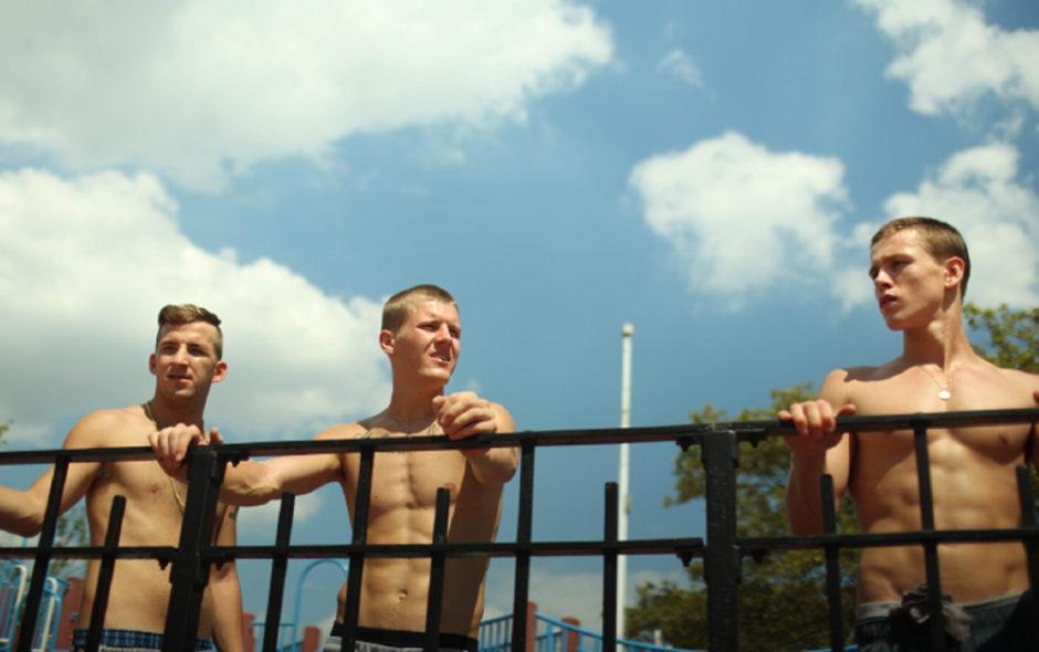 Press still of the movie Beach Rats. Nick (Frank Hakaj), Alexei (David Ivanov) and Frankie (Harris Dickinson) shirtless leaning on railing outside.